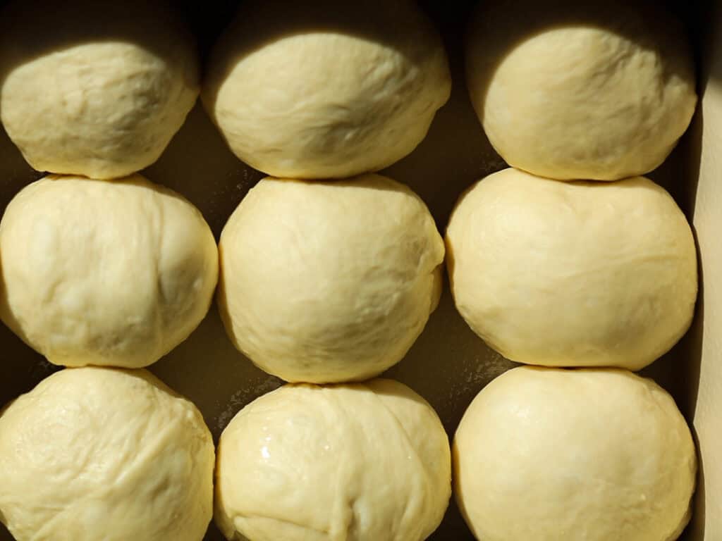 Eight round balls of dough arranged in rows, resting on a surface. The doughs surface is smooth and slightly shiny, illuminated by natural light.